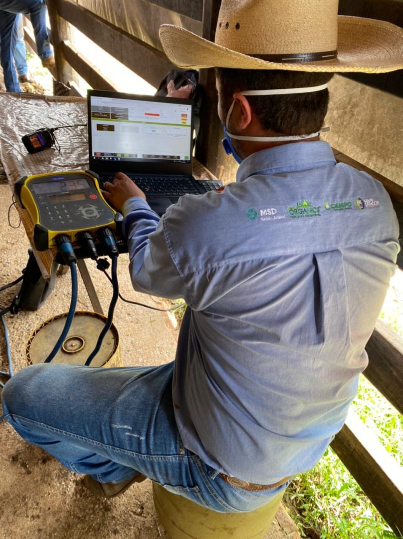 Na imagem, temos o retrato de um homem em frente a um notebook configurando sistema de gestão de pecuária de corte em um curral. Ao lado do notebook, vemos um equipamento. Homem visto de costas está de camisa azul, calça jeans e chapéu.
