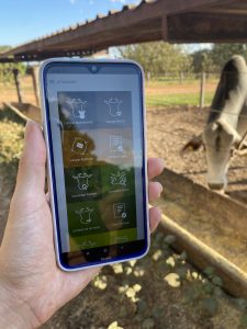 Na imagem, temos uma mão feminina segurando um celular na tela do iRancho. Ao fundo, em um pasto, vemos um animal se alimentando. Foto ilustra matérias de ESG no agro.